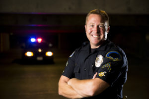 A smiling police officer with his patrol car in the background.